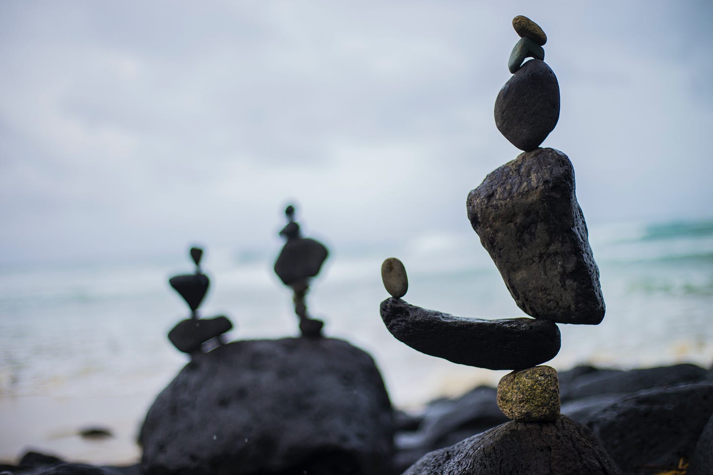 3 pieces of intricately stacked rock art, in black rocks, on a cloudy beach that is blurred in the background 