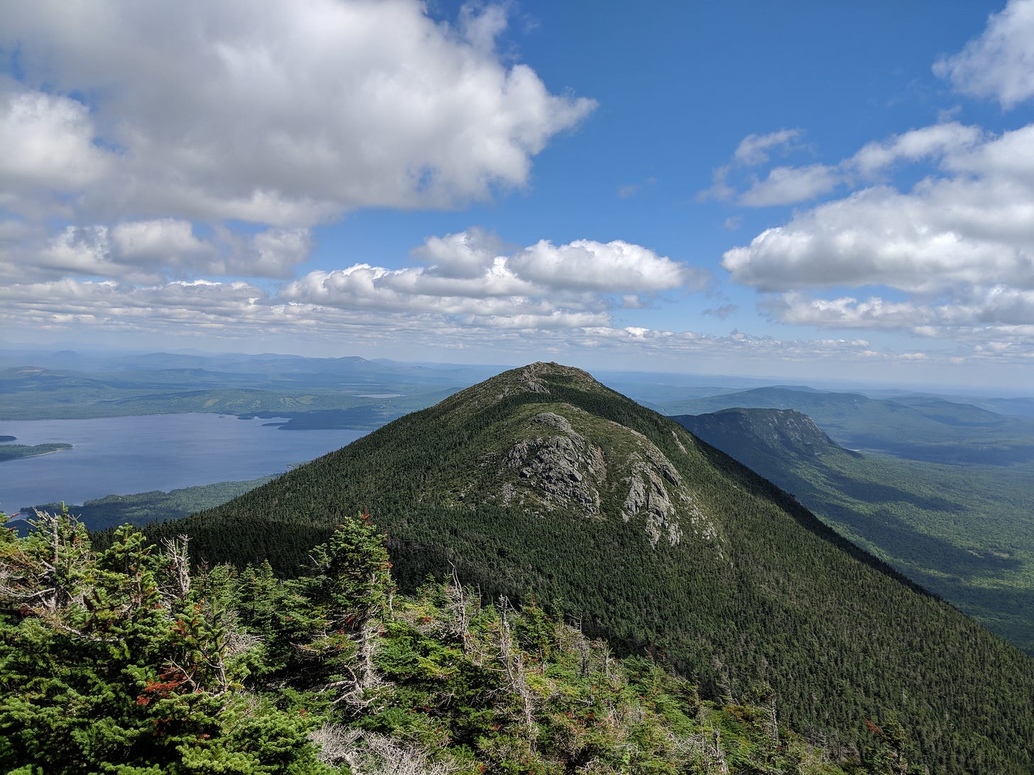 Photo of Avery Peak in the Bigelow Mountains of Maine, taken while standing on West Peak. The sky is blue and full of puffy clouds, and Flagstaff Lake sits to the left behind Avery Peak.