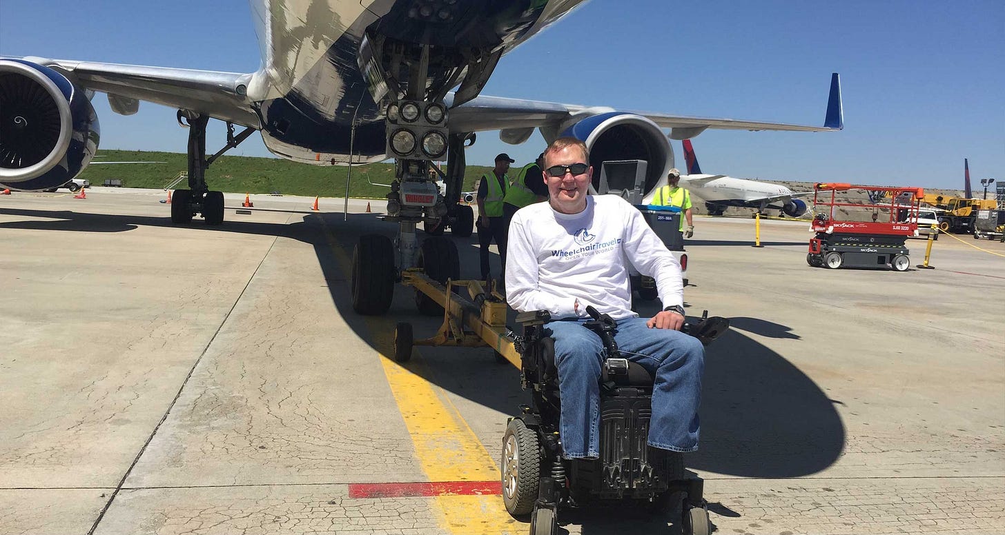 John seated in his wheelchair in front of an airplane on the tarmac at an airport hangar.