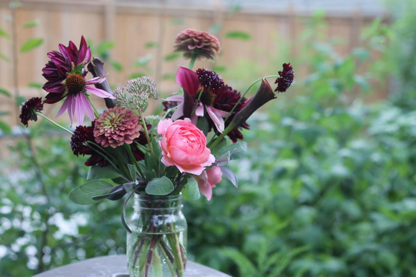 A bouquet of pink and purple flowers sits in a mason jar on a table outside.