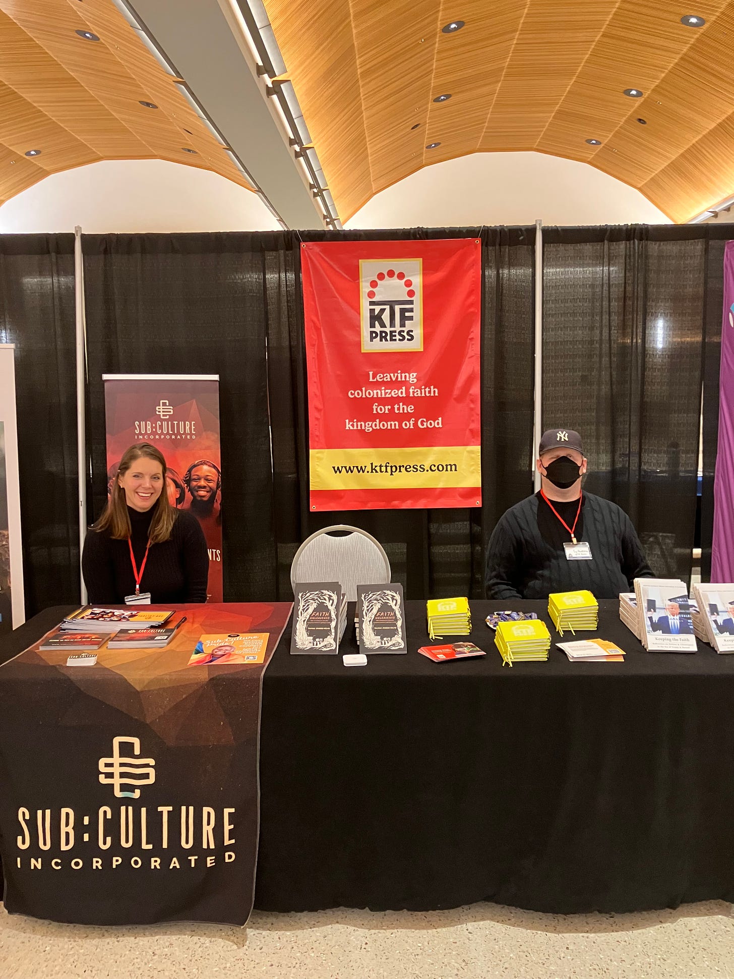 A photo of Sy standing at KTF’s booth at the Evolving Faith Conference. The booth is shared with Sub:Culture, Inc. Tamice’s nonprofit. A tall White woman is also at the booth with Sy standing on the side of the table dedicated to Sub:Culture. KTF’s books and other materials are on the table, and a large sign for KTF is hung behind Sy.