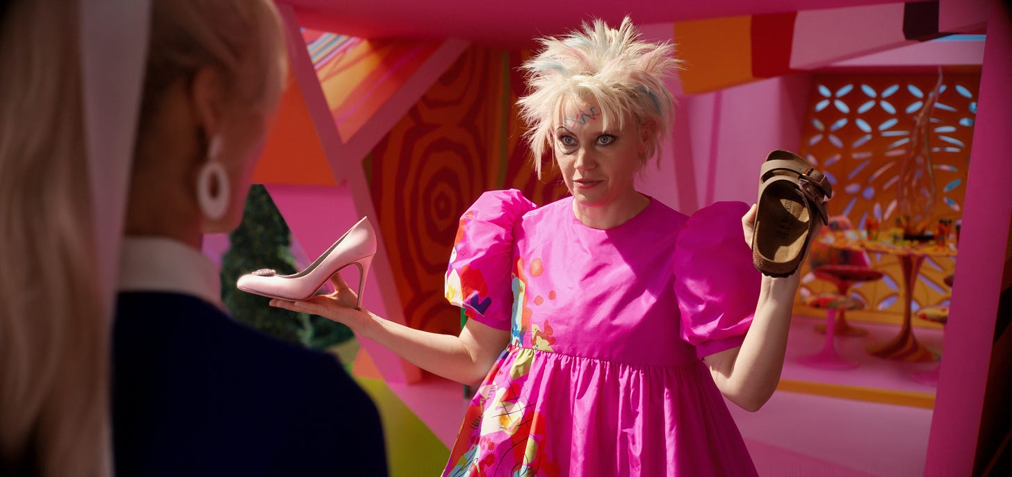 Margot Robbie as Stereotypical Barbie (left, from behind) and Kate McKinnon as Weird Barbie (right) in BARBIE. Weird Barbie is holding a pink heeled shoe in her right hand and a Birkenstock sandal in her left hand.