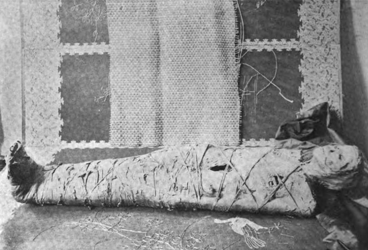 A wrapped mummy lies on either a table or the floor. It looks decayed and ancient.