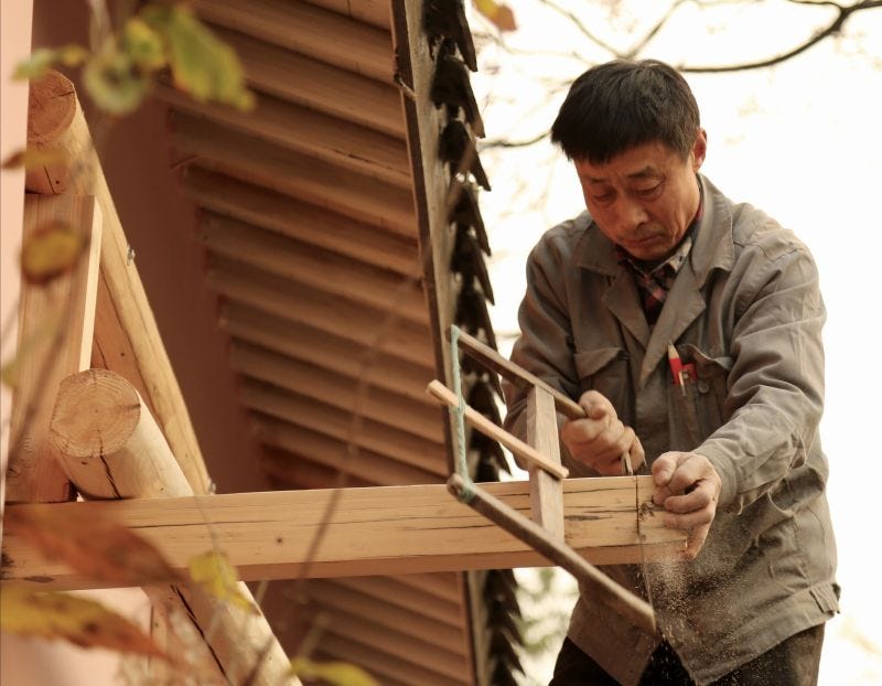 A man uses a manual handsaw to cut a beam on a wooden structure that appears to be underconstruction.