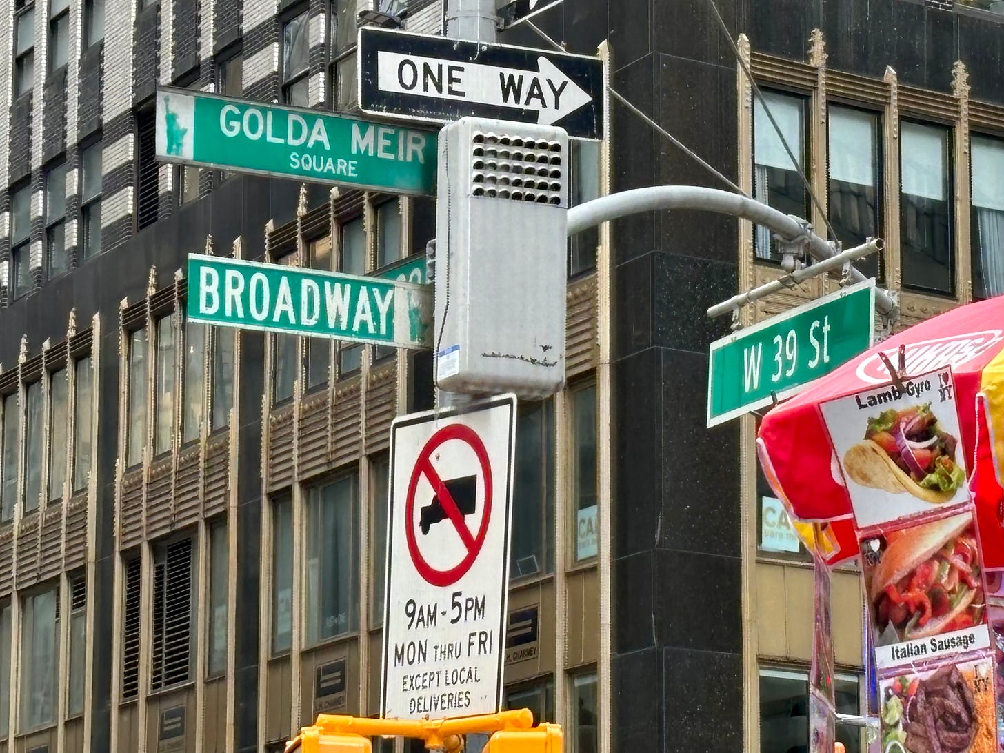 Three street signs at Broadway and W 39th St giving the intersection and markign Golda Meir Square. There is also a food cart with a sign for a lamb gyro.