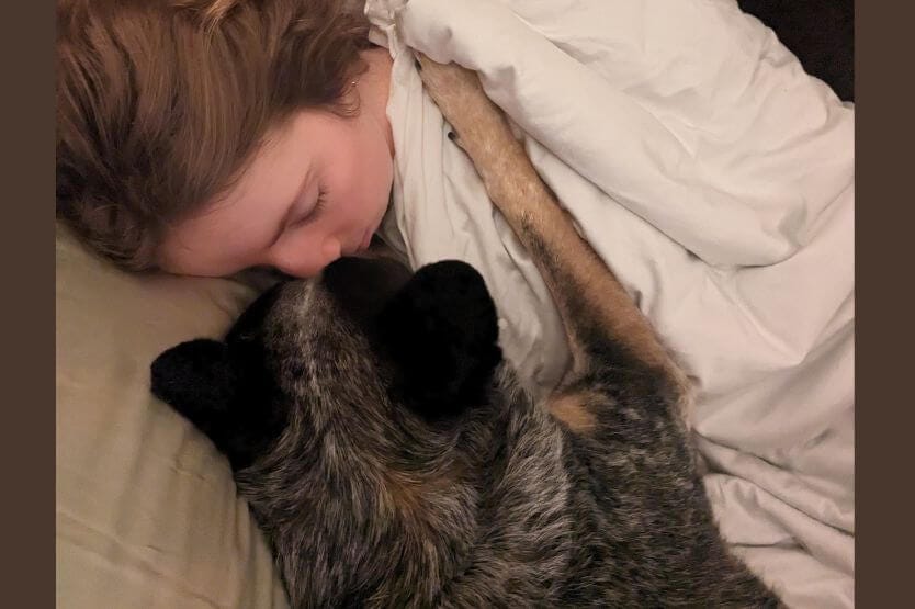 A close up photo of Haley the young woman and Scout the blue heeler snuggling in bed, foreheads pressed together