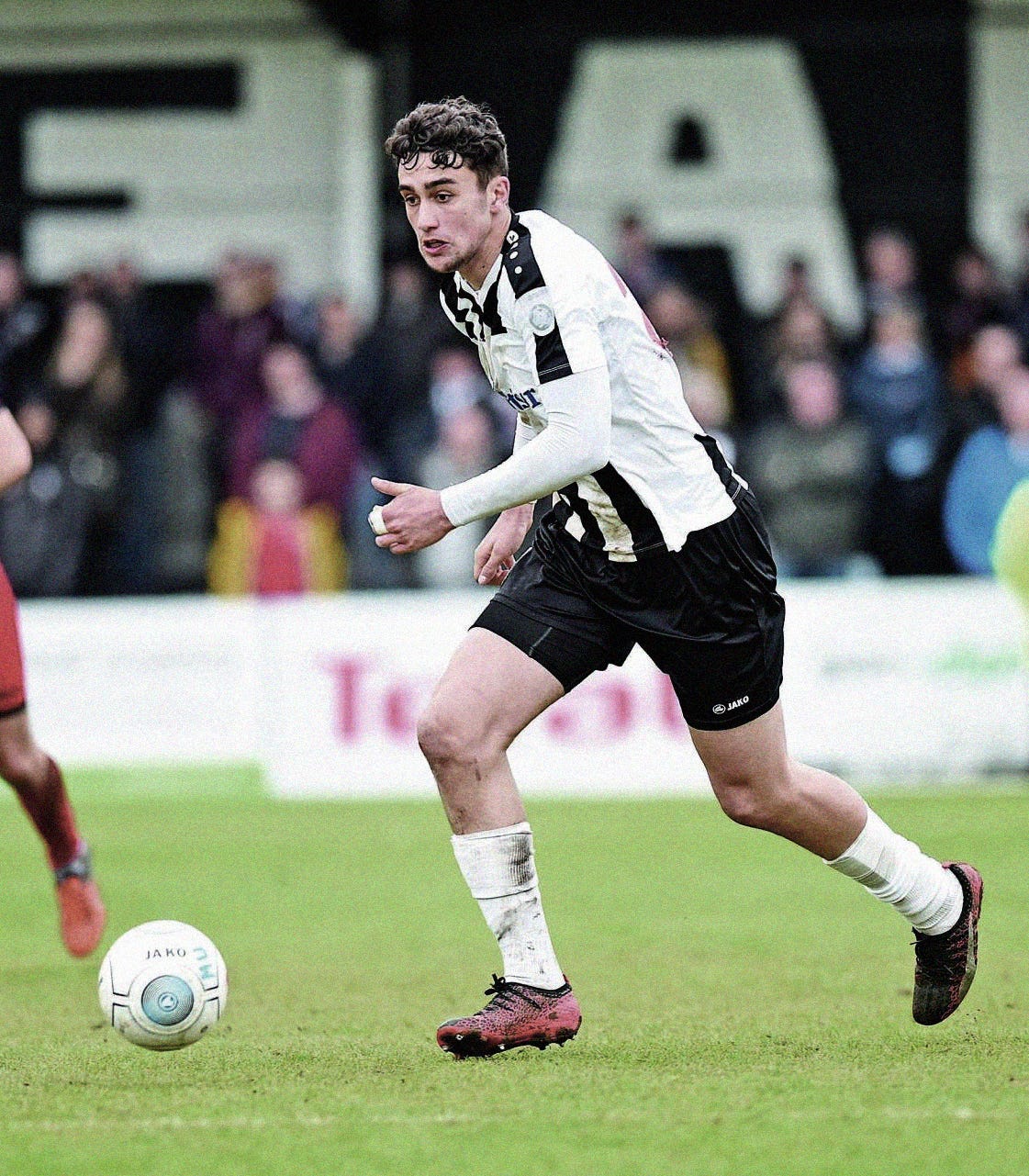 A photo of Max Kilman playing for Maidenhead United in the National League. He's wearing a black-and-white striped shirt with black shorts, white socks, and black boots. He's running with the ball with a blurred crowd scene in the background.