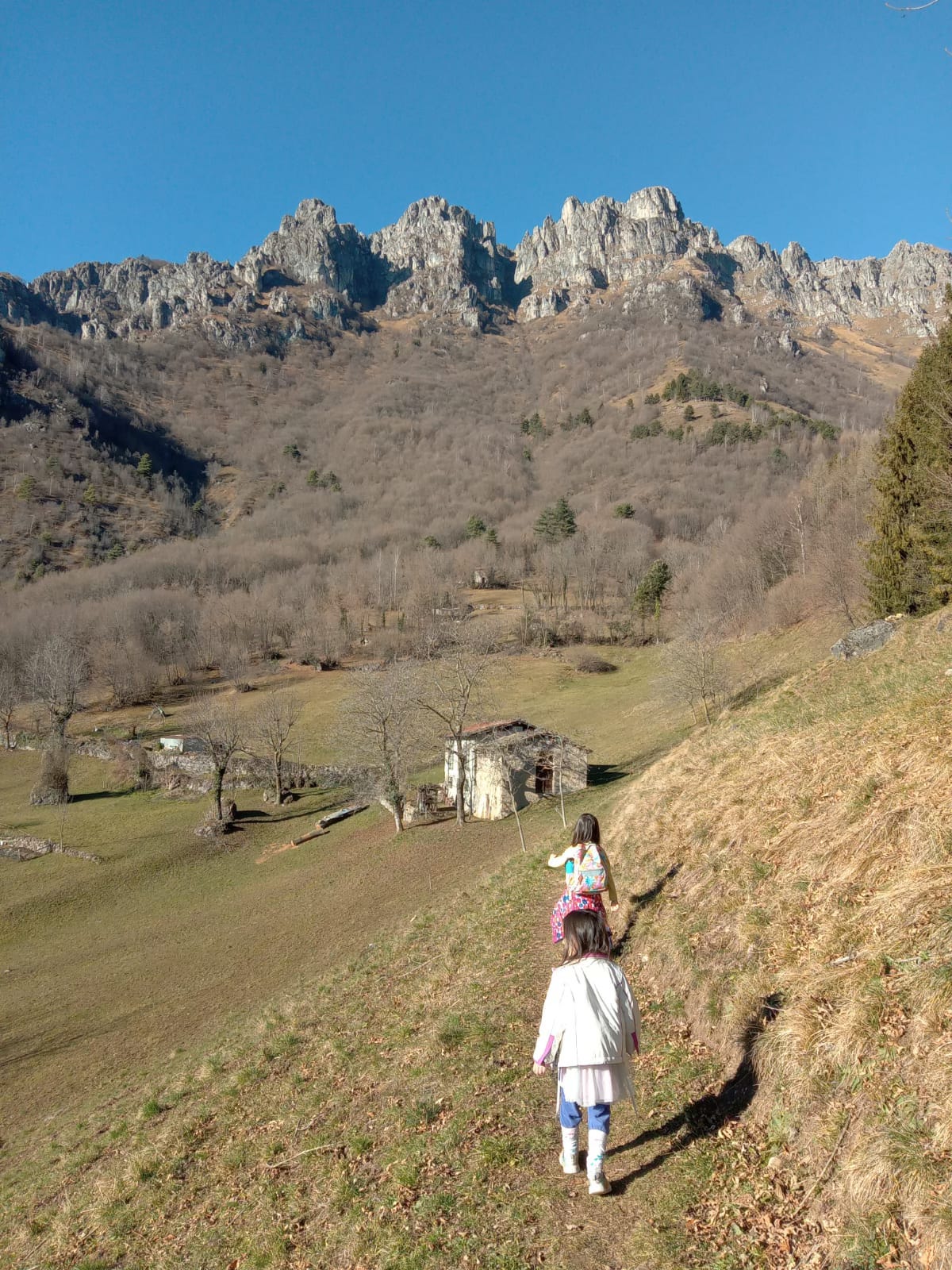 Un bambino e una bambina camminano su un sentiero di montagna. Sullo sfondo si stagliano delle vette rocciose e un cielo nitido e azzurro intenso.