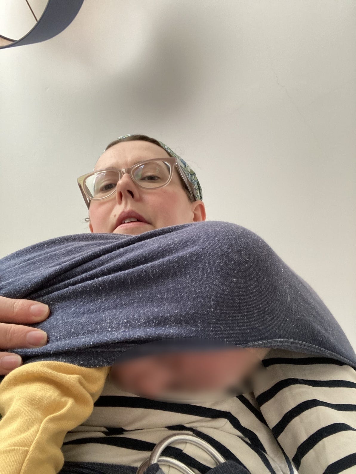 Woman in glasses looking down to the camera. She has a baby in a stretchy wrap but the baby has worked his way down and his face is pointing out of the wrap.