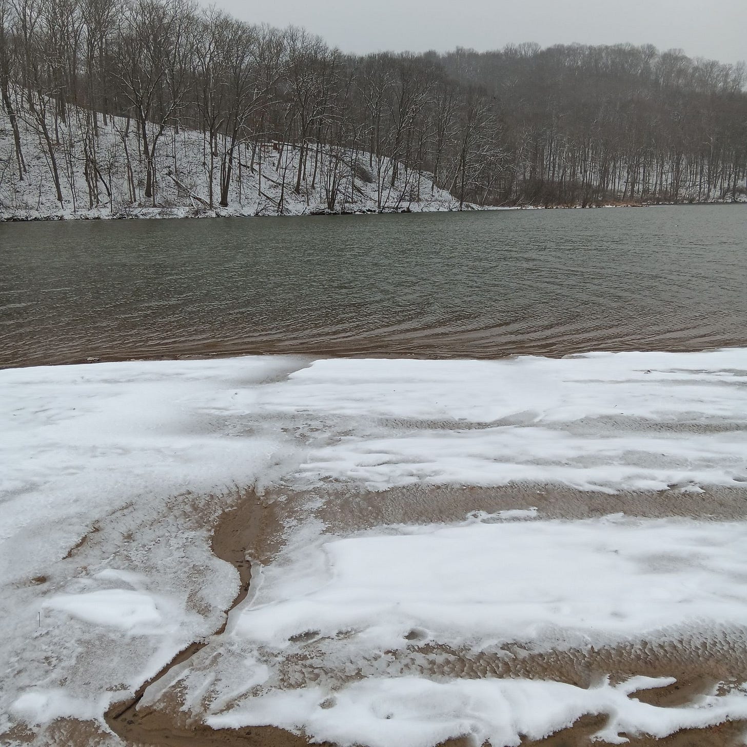 The lake at Raccoon Creek, rippling in the winter wind. The beach is covered in snow that's streaked with patterns where it got wet. There are bare trees across the lake. 