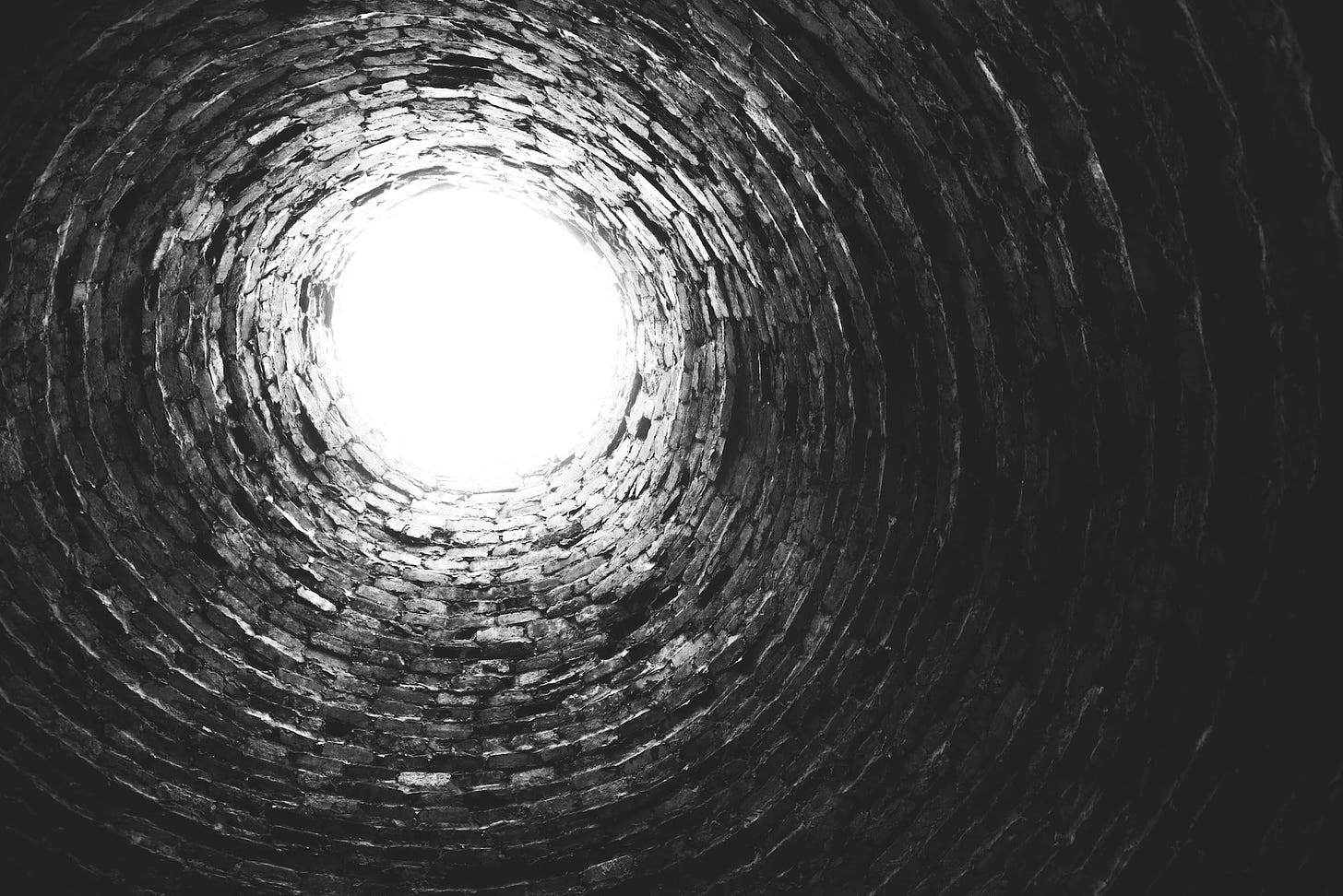 A black and white photograph looking upwards at the round circle of white sky-light from inside a brick-lined well.