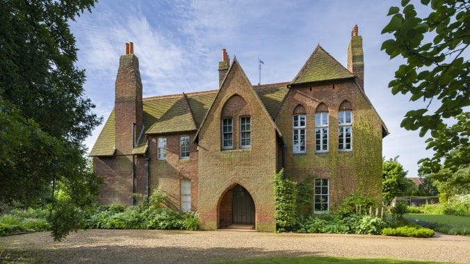 An image of the imposing red brick facade of Red House in London showing its north front with arched entrance way and windows surrounded by gardens