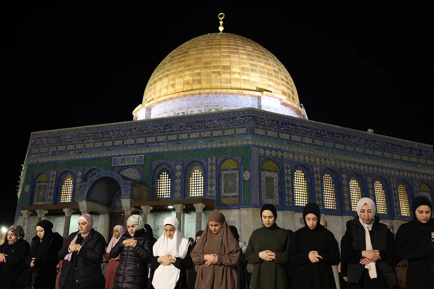 Palestinian Muslim women stand in a row in front of Al-Aqsa Mosque in Jerusalem with their hands folded and their heads bowed in prayer. The gold Dome of the Rock stretches into the night sky behind the women.