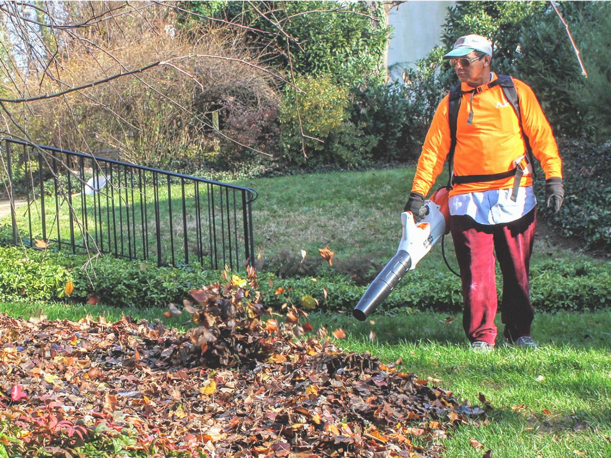 A person wearing an orange top and red pants blowing leaves with a battery powered leaf blower