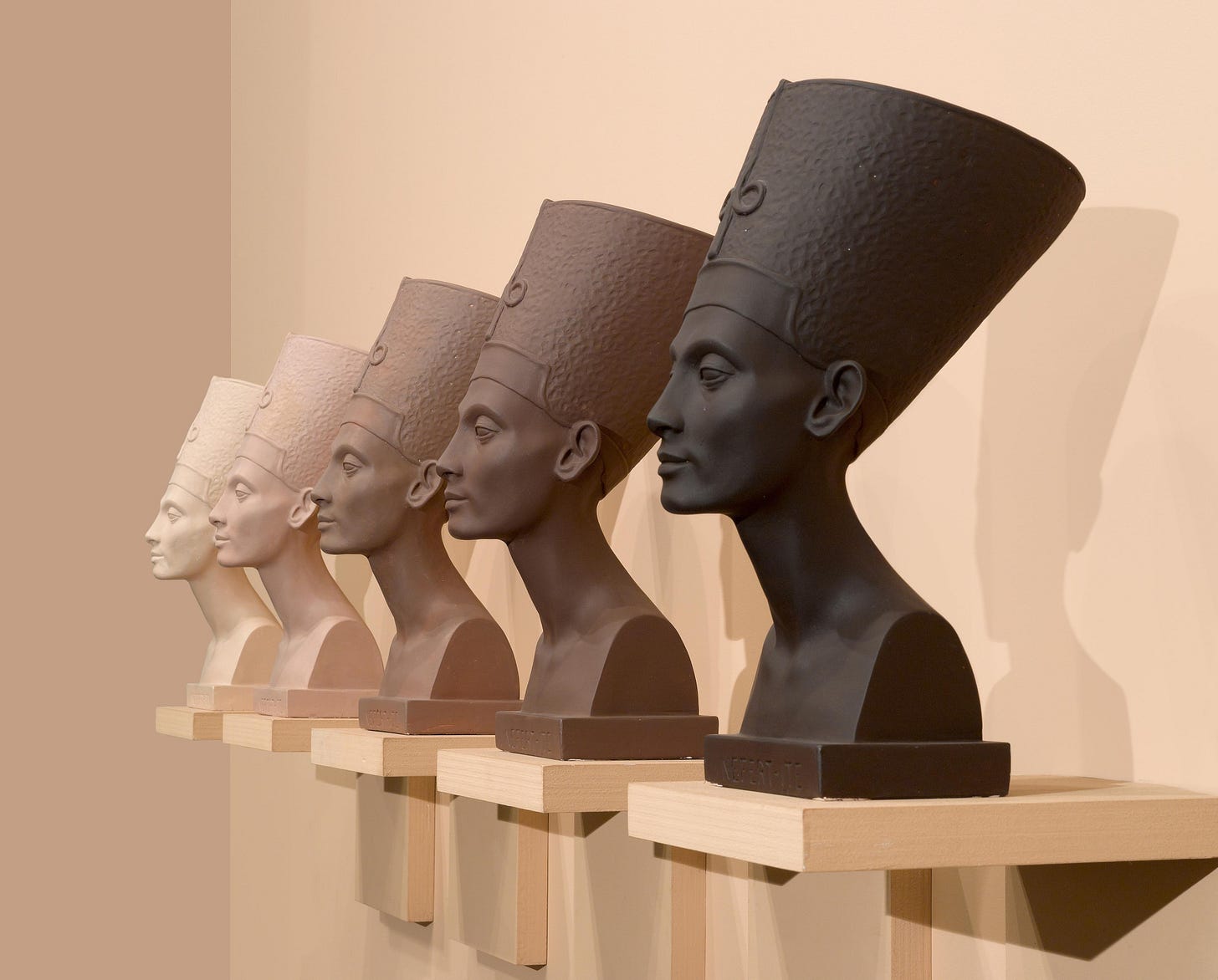  A row of busts depicting the famous bust of Nefertiti, each a different shade of brown ranging from almost-black to white-ish beige, set against a peach-colored background.