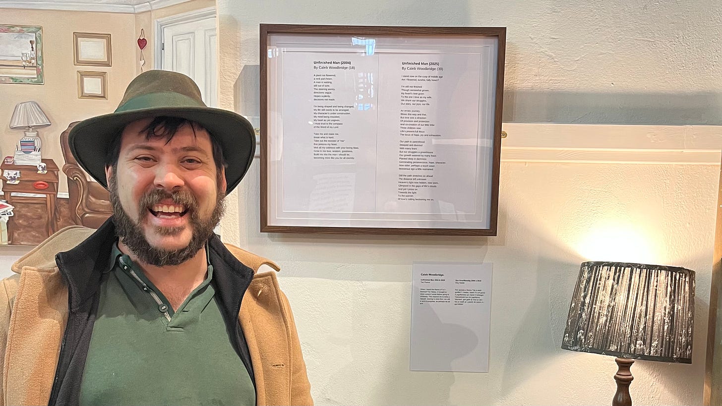 A bearded man in green hat (me!) standing in an art exhibition in front of his poems mounted on the wall