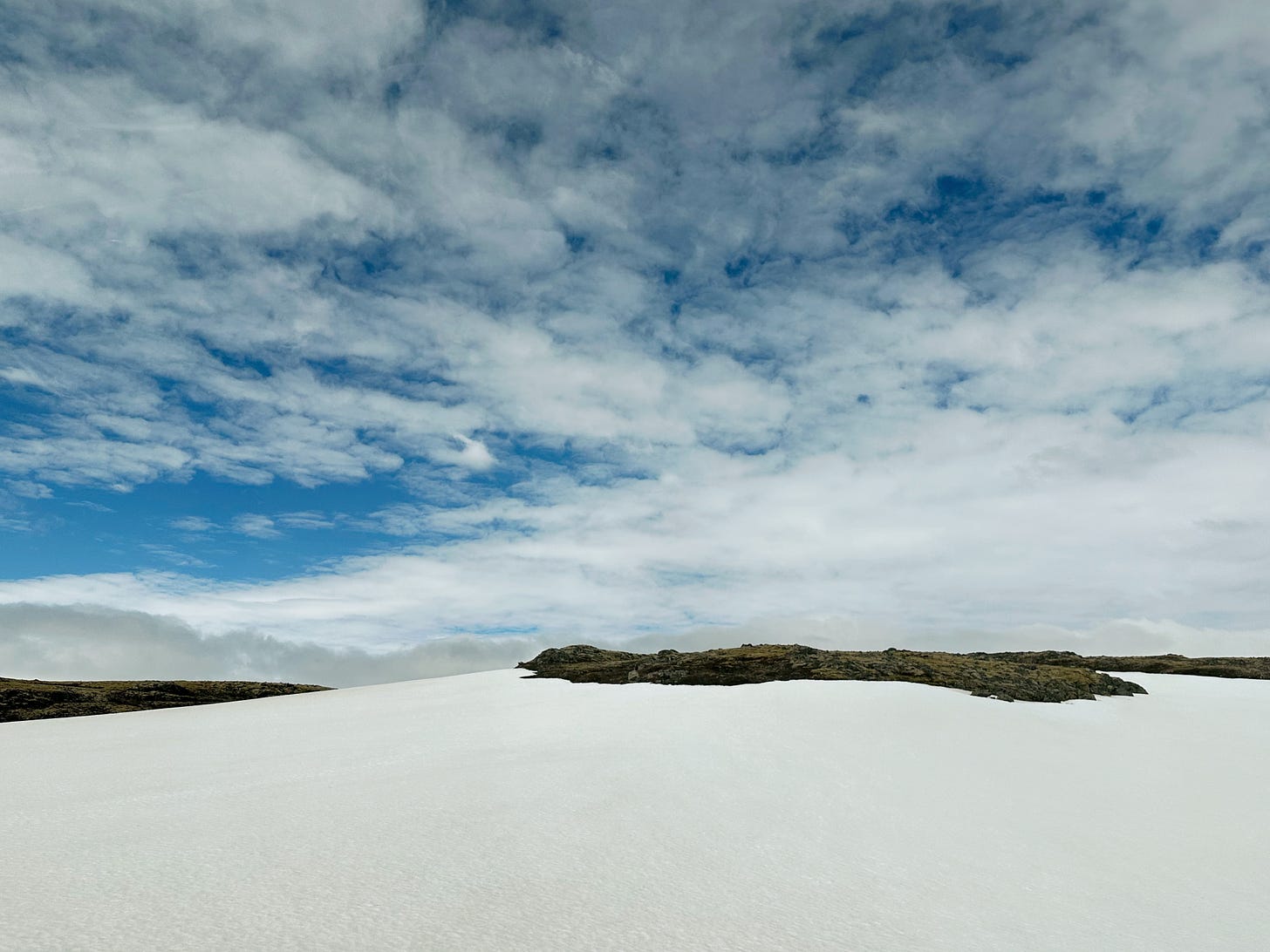 Snow filled mountain pass with small patches of exposed lava rock peeking through the snow.  The sky is bright blue with puffy, white clouds that blend into the snow on the ground.