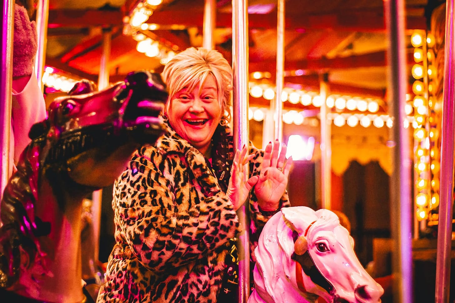 A joyful woman with short hair, wearing a leopard print jacket, is smiling broadly and waving towards the camera. She is on a carousel at a fair, surrounded by lights that create a warm, vibrant atmosphere. A white carousel horse with pink highlights and a golden mane is in the foreground, with the pole structure of the carousel providing a bright, festive background. Her expression and the setting evoke a sense of fun and nostalgia.