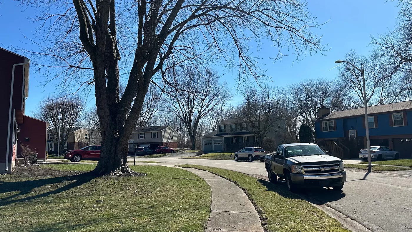 View from down the sidewalk of a typical American 1970's suburban subdivision. The road curves gently to the left and there are a few cars parked in the street at the curbs on both sides. There is no traffic. The trees are bare because it is early March in Ohio and the sky is crystal blue.