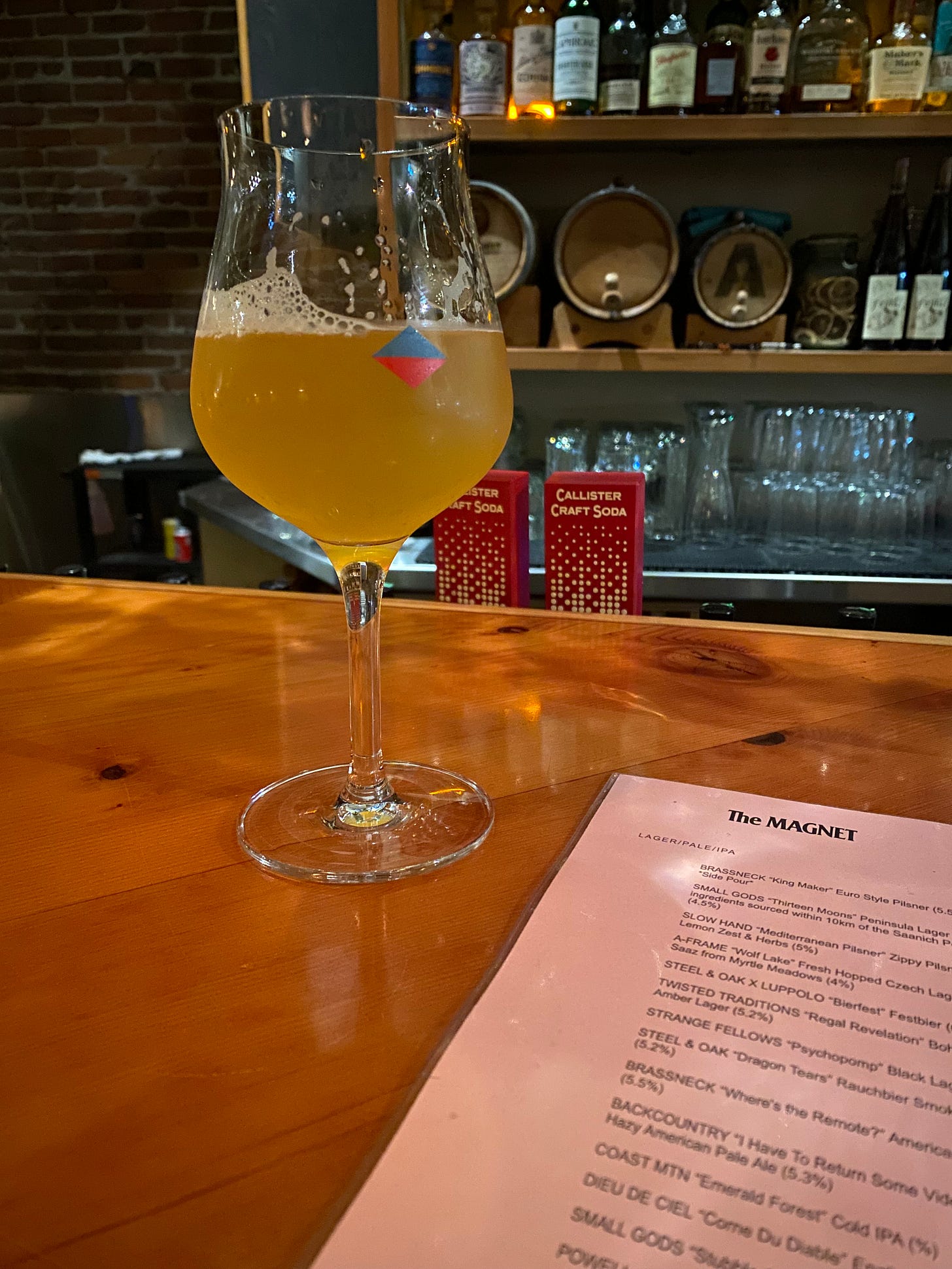 A tulip glass of IPA on top of the bar at The Magnet; the beer list is visible in the bottom right corner. In the background bottles of hard liquor and empty glasses of various sizes can be seen on the bar shelves.