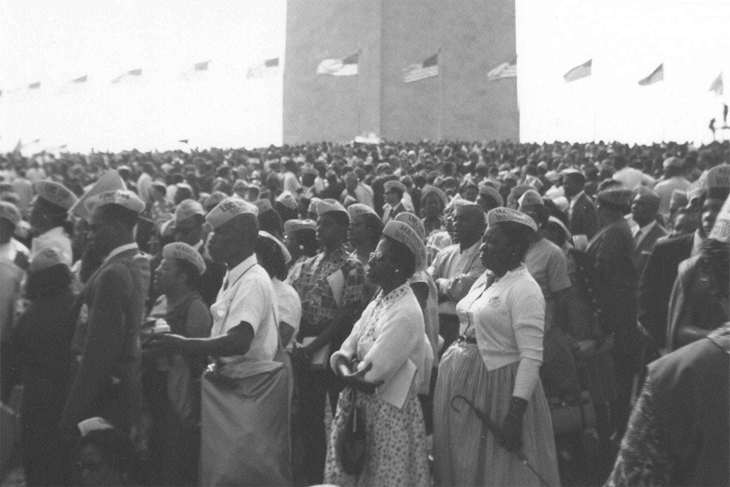 A black-and-white photo of the thousands gathered at the march watching a speaker against a backdrop of American flags. 