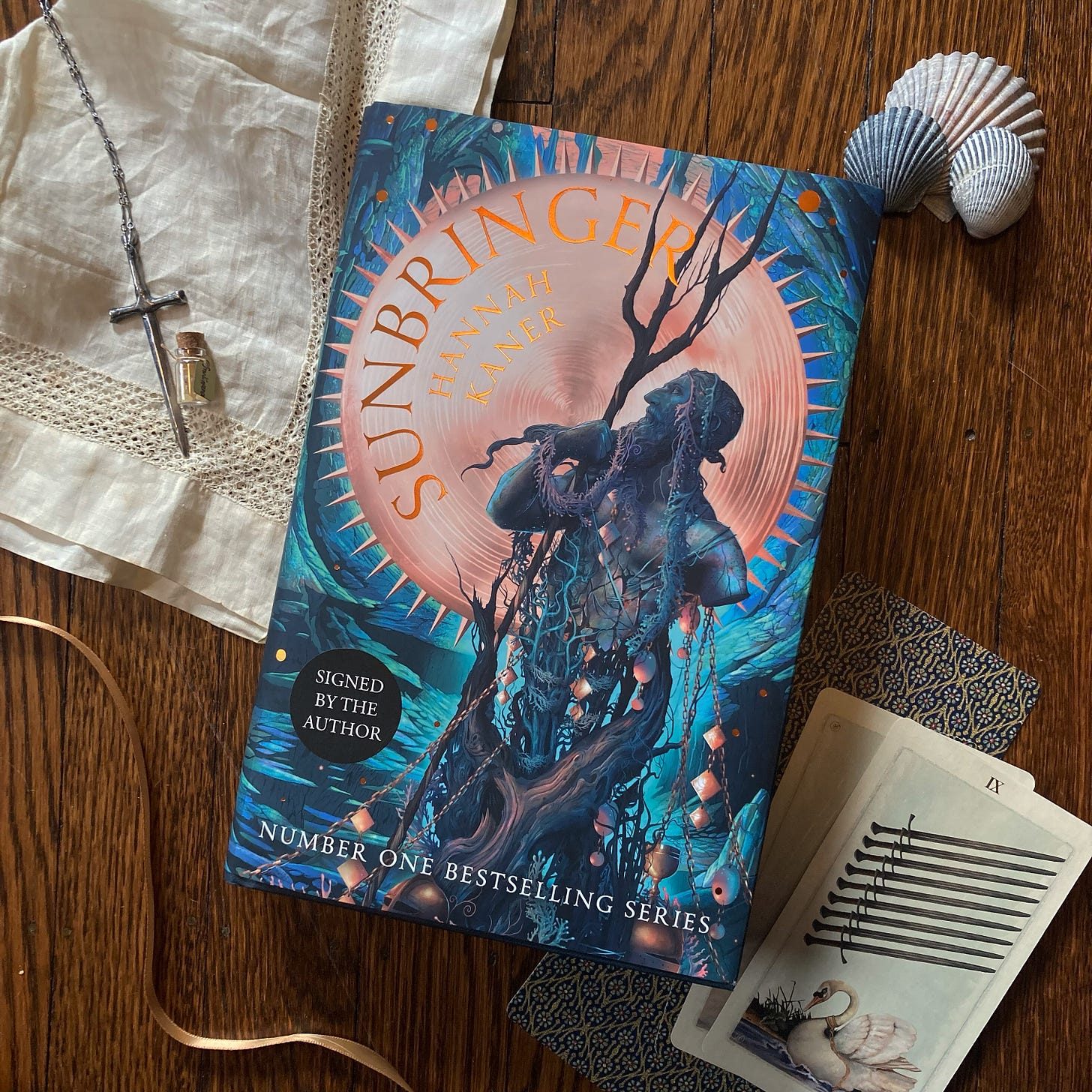 hardcover copy of Sunbringer in a flat lay featuring tarot cards, a golden ribbon, a sword necklace, vintage cream handkerchief, and shells
