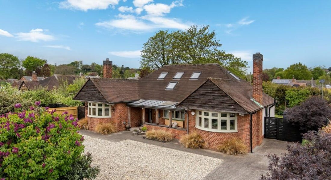 A brown and brick dormer bungalow with a driveaway in front of it and colourful bushes and greenery around it