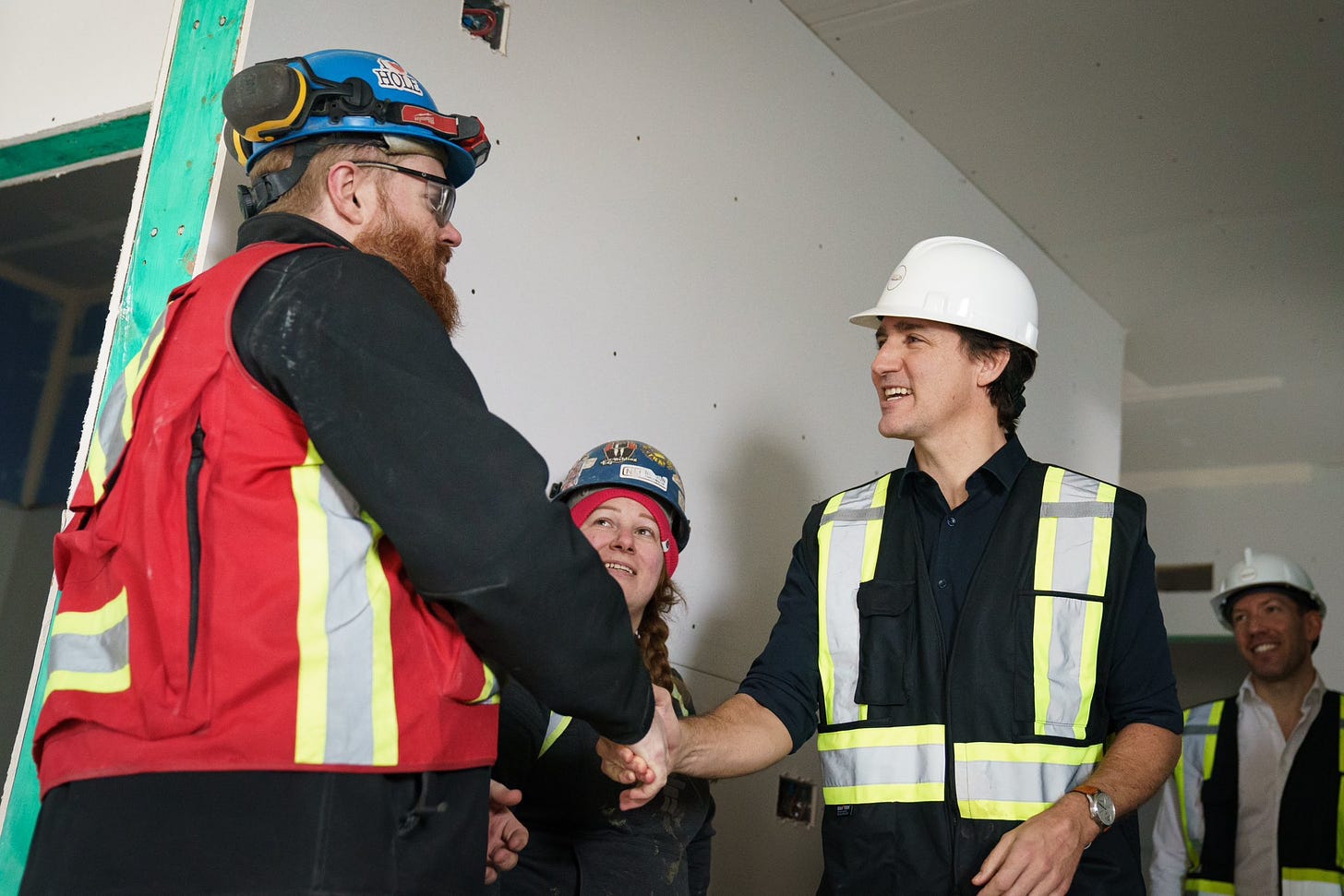 Prime Minister Justin Trudeau is standing and shaking hands with a man, who is standing in front of him. A woman and man are standing behind them. Everyone is wearing safety vests and hard hats, and is smiling.