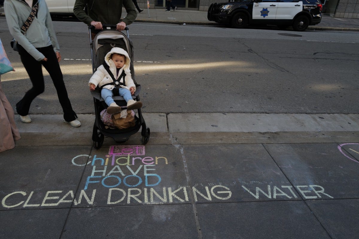 A child in a stroller crosses over a chalked message concerning a child’s right to food and clean water outside of the Israeli Consulate.