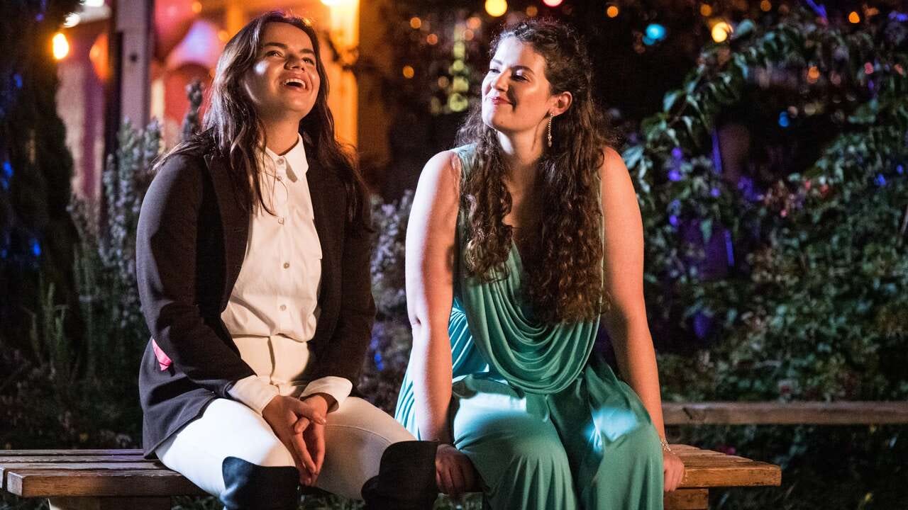 In an outdoor night scene lit with colorful garden lights, two young people sit side-by-side on a wooden bench. The young person on the left has long brown hair and is wearing a white-and-black horseriding outfit as they look up smiling. The young woman on the right has long curly brown hair and is wearing a drapey mint green formal dress. She's smiling as she looks over at the person next to her.