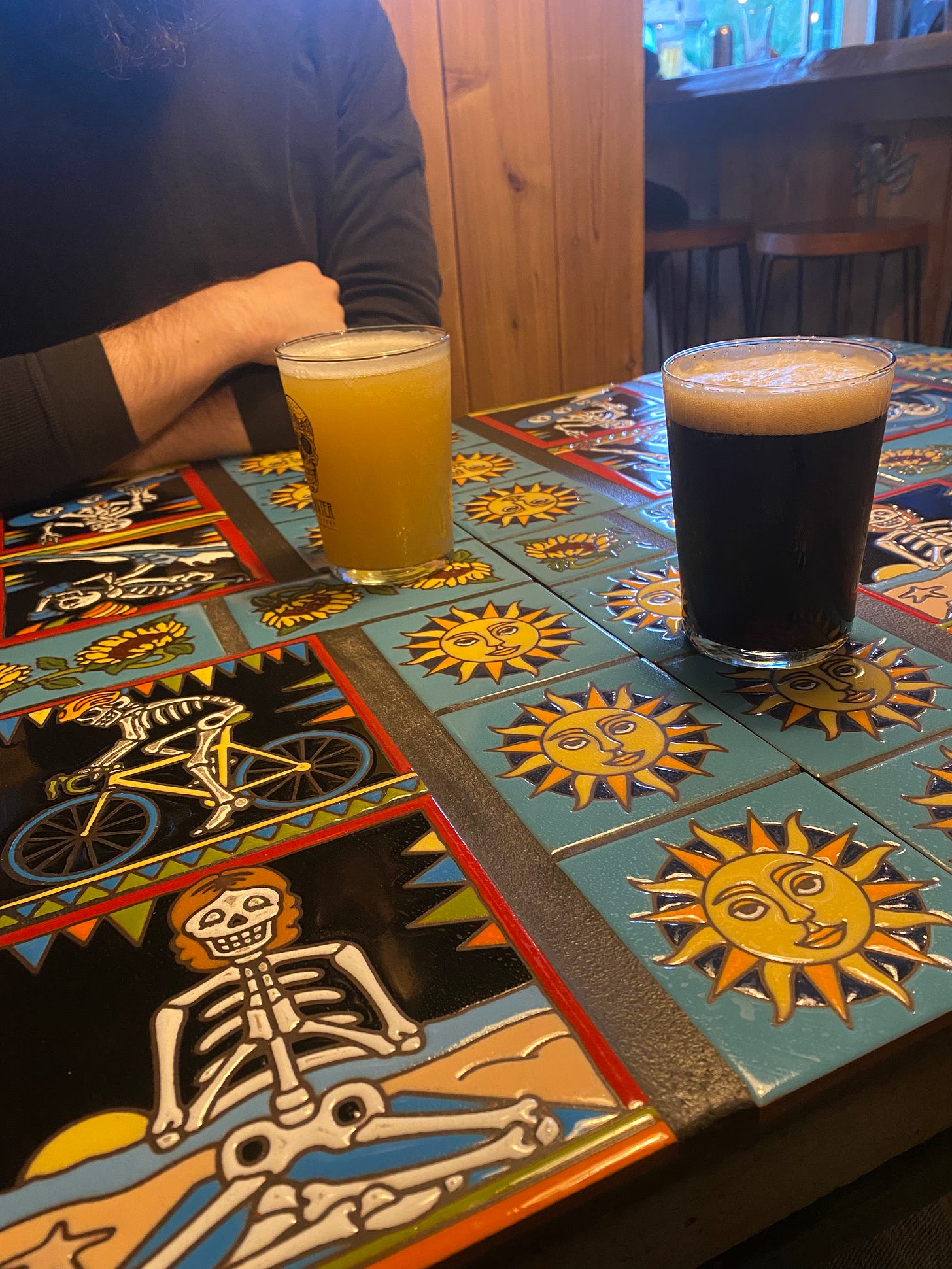 A tiled table with two glasses of beer on it, dark in the foreground and light in the background, where Jeff's sitting. The tiles are a mix of art deco-style suns with faces, and skeletons doing North Van activities like biking and skiing and yoga.