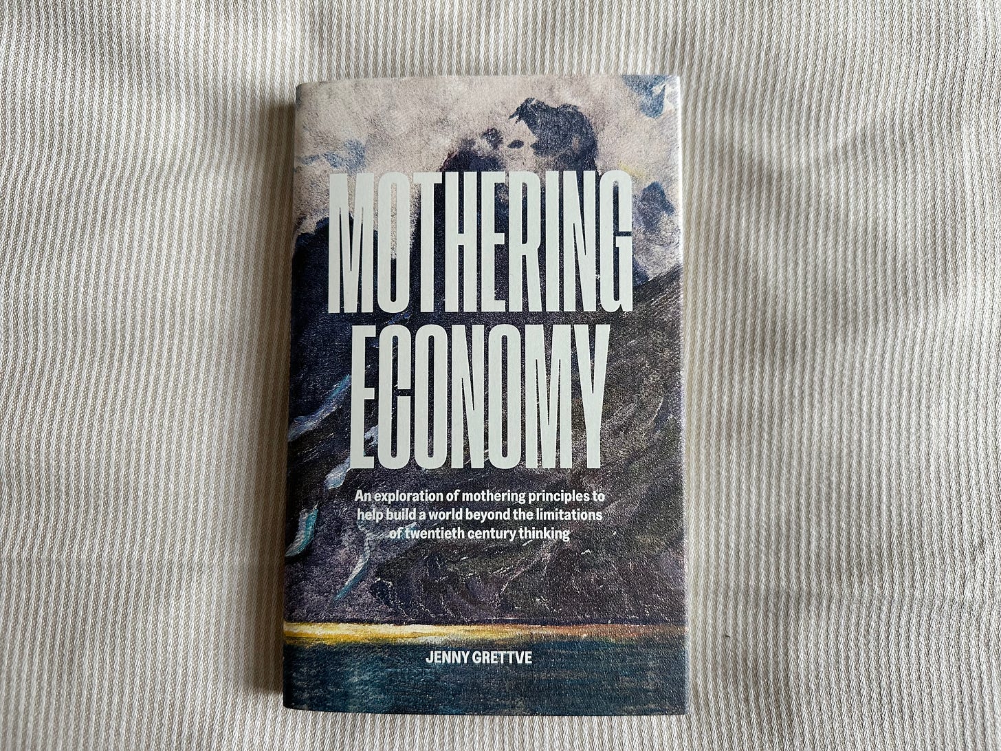 Picture of a book titled "Mothering Economy" by Jenny Grettve, on a fine striped beige and white duvet. The cover shows detail of a landscape painting with the title “MOTHERING ECONOMY” in large san-serif white font.