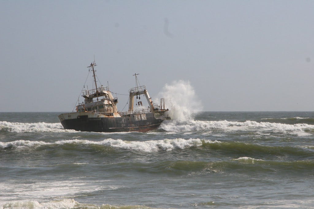 A rusted abandones shipwreck off the coast of Namibia is battered by waves on an overcast day.