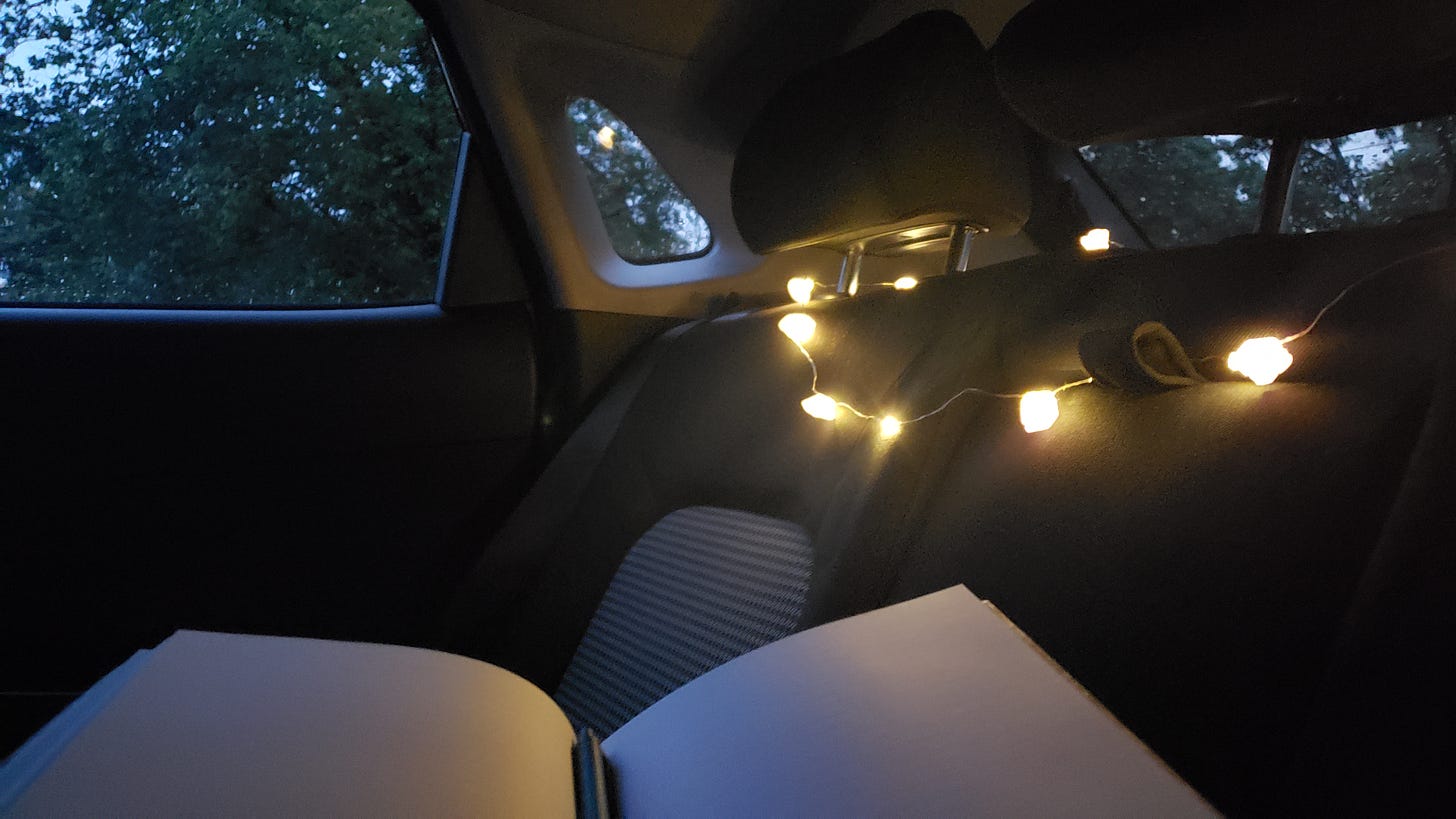 Perspective of someone sitting in the backseat of a car with open notebook on their lap. Large purple-white string lights are draped between the headrests hanging over the backseat. In the left corner a bit of twilight blue sky and green leaves can be seen through the car window. 