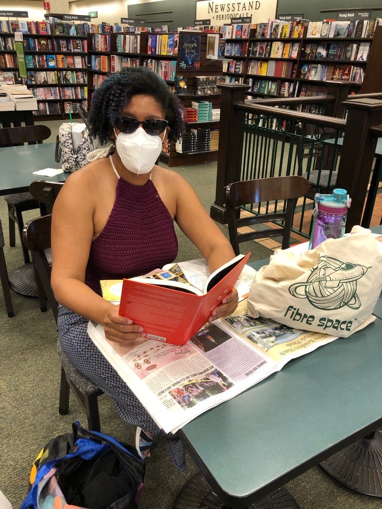 Kristen is facing the camera, seated at a table in the Potomac Yard Barnes and Noble. She is holding a book and she has a bag of yarn and a water bottle on the table. She is masked and has on sunglasses.
