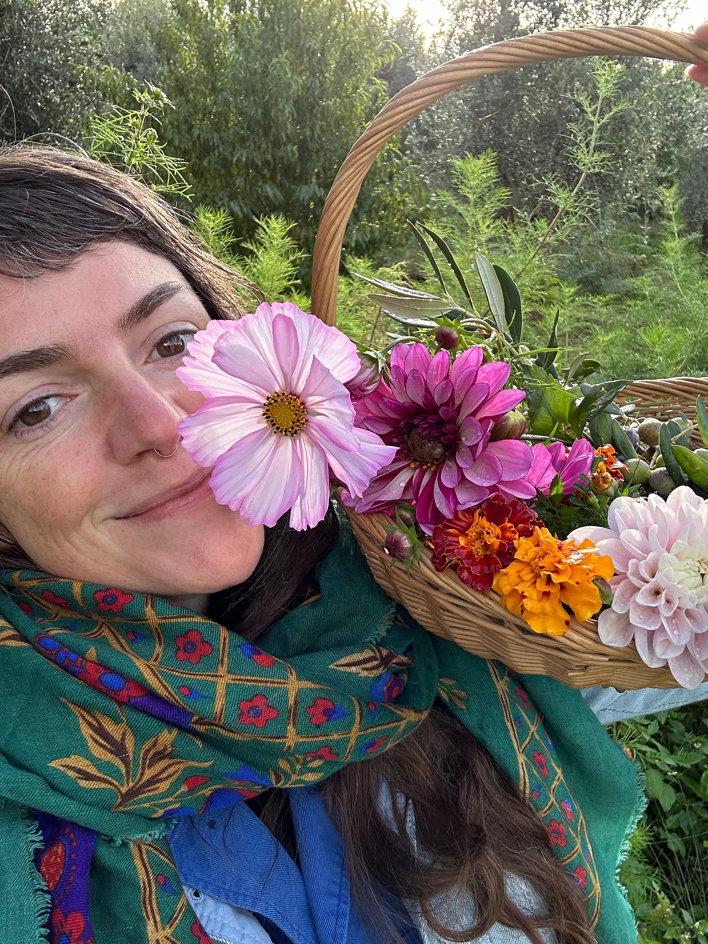 Me smiling at the camera holding a basket up filled with dahlias, cosmos and marigolds. Trees are behind me. 