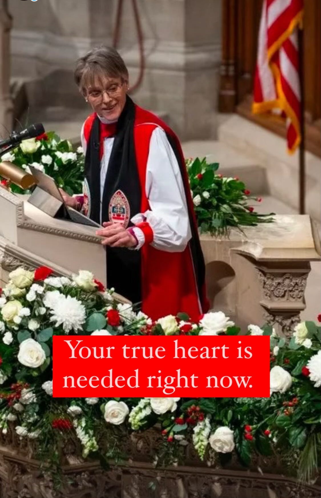 A color photograph of Bishop Mariann Edgar Budde standing at the pulpit at the National Cathedral. She is a white woman with ear-length grey-brown hair, wearing Bishop’s vestments in white, black, and vivid red, and she is smiling as she turns her head to look downward. Below the pulpit is an abundant array of greenery and white and red flowers. Over the image are the words “Your true heart is needed right now.” 