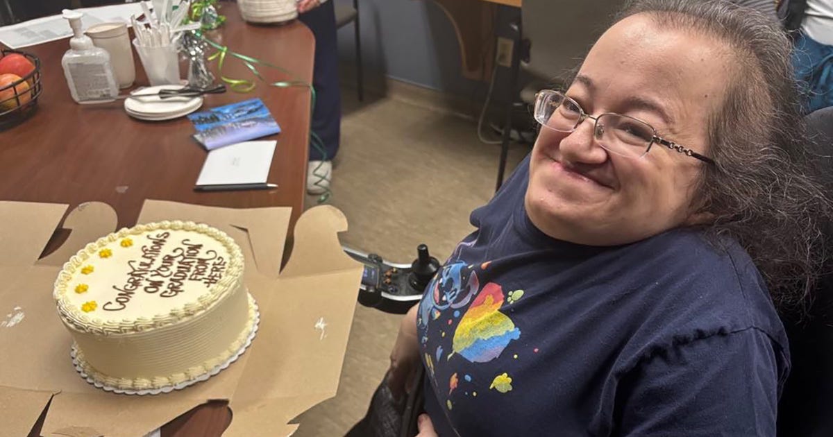 Athena sitting and grinning in her power wheelchair in front of a cake that reads "Congratulations on Your Graduation from Here"