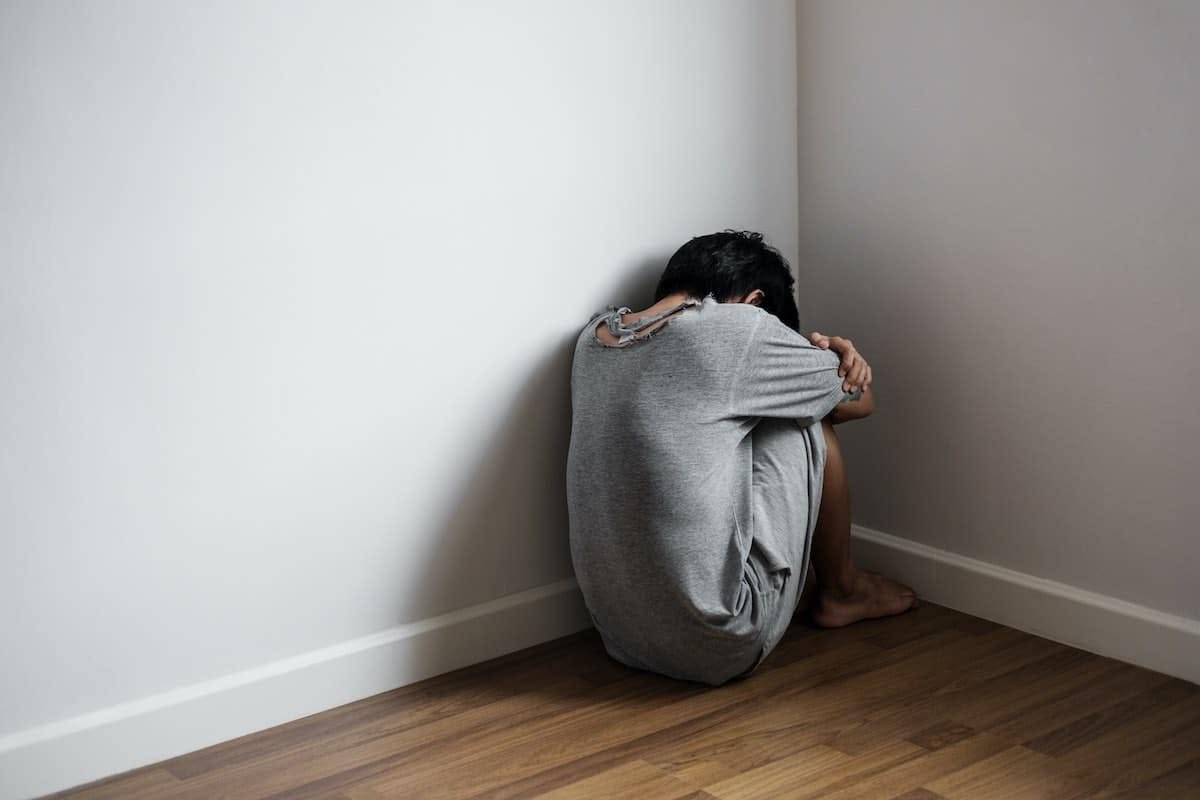 Depressed person, knees drawn to chest, arms wrapped around them, head tucked, facing a corner.