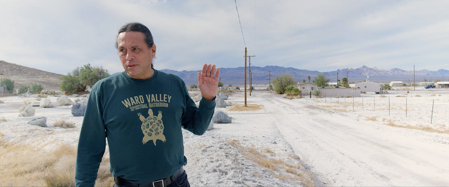 Ian Zabarte, who has black hair in a ponytail, wears a blue shirt against a desert backdrop