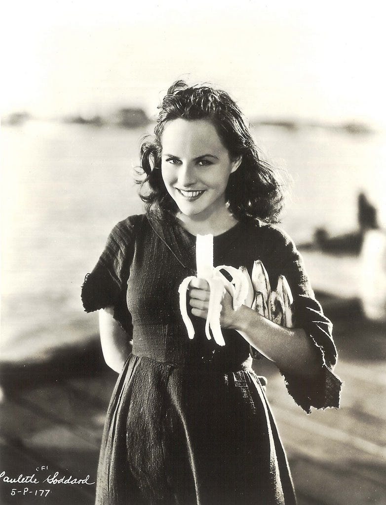Paulette Goddard, actor, in black and white still from 1936 film Modern Times. She's an urchin who'se holding some bananas stolen from the cosk and smiling at the camera