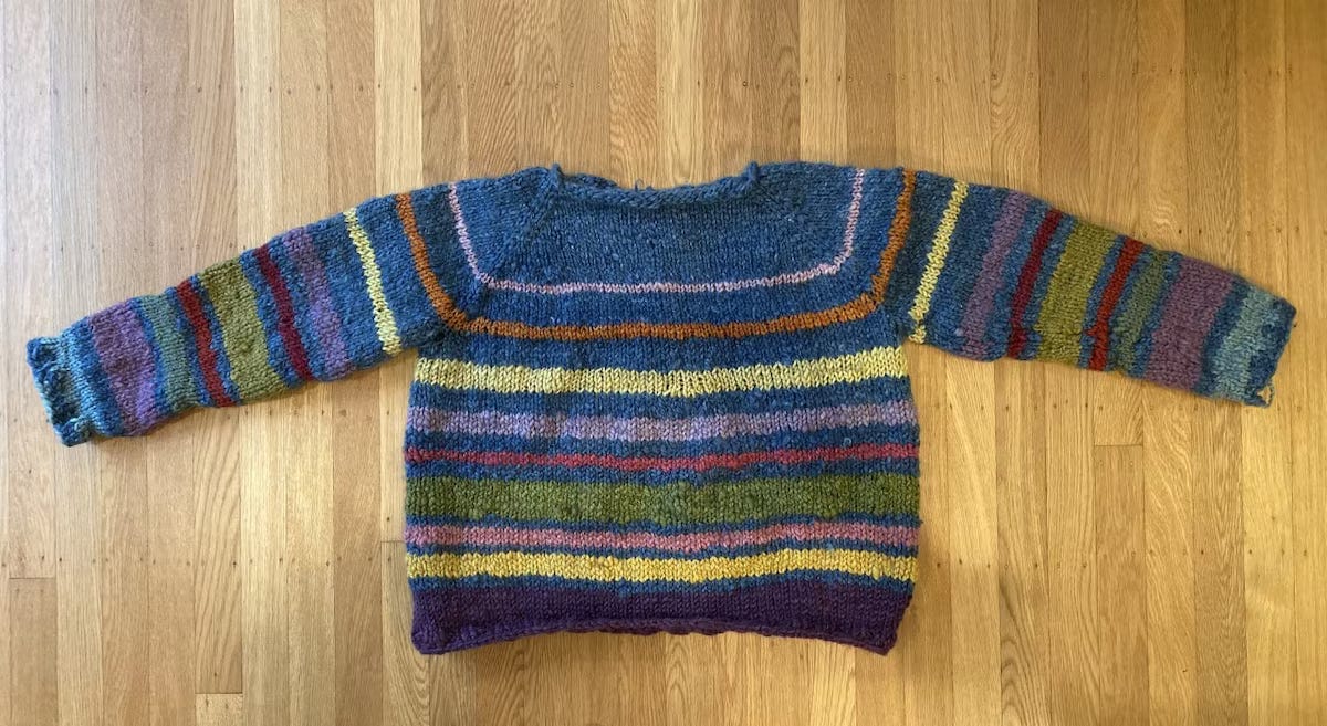 A hand-knit sweater with many colored stripes and a mostly blue top, laying flat on a wooden floor.