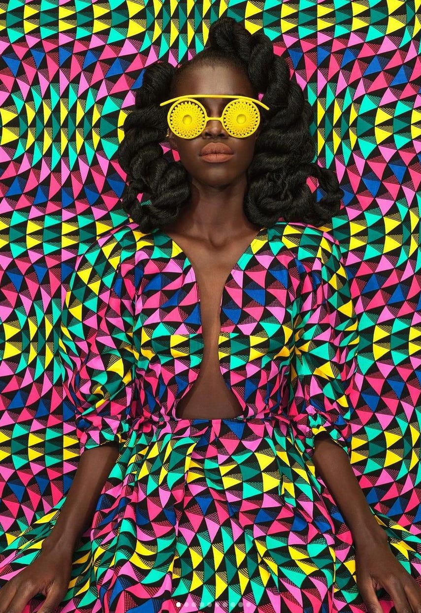A photo of a Kenyan woman wearing yellow sunglasses and a colorful patterned dress which matches the background behind her