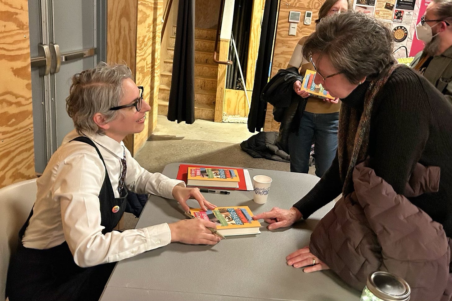 Left: I'm sitting behind a table with two copies of Endpapers in front of me. Standing across from me (right) is a woman leaning on the table, telling me who to sign her book copy for. I'm smiling at her. Behind her more people hang out and wait in line.