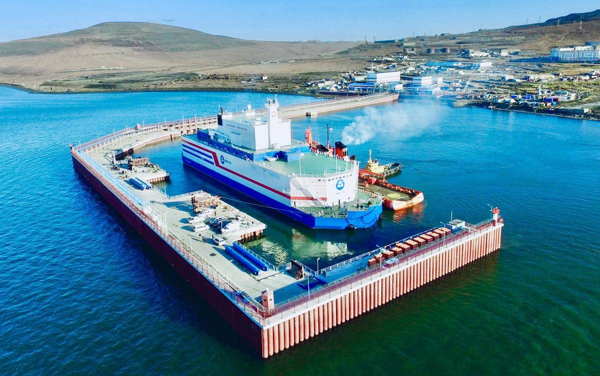 A floating nuclear power plant docked at a harbor, surrounded by calm water, with hills and buildings in the background.