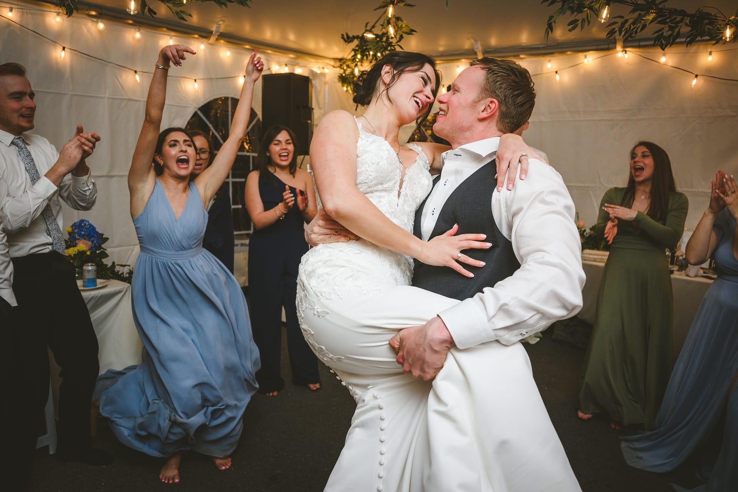 A groom lifts his bride while wedding guests celebrate behind them