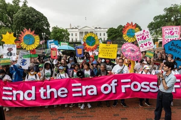 Dozens of people carrying brightly colored signs and a large pink banner that reads “End the Era of Fossil Fuels” are gathered in front of the White House.