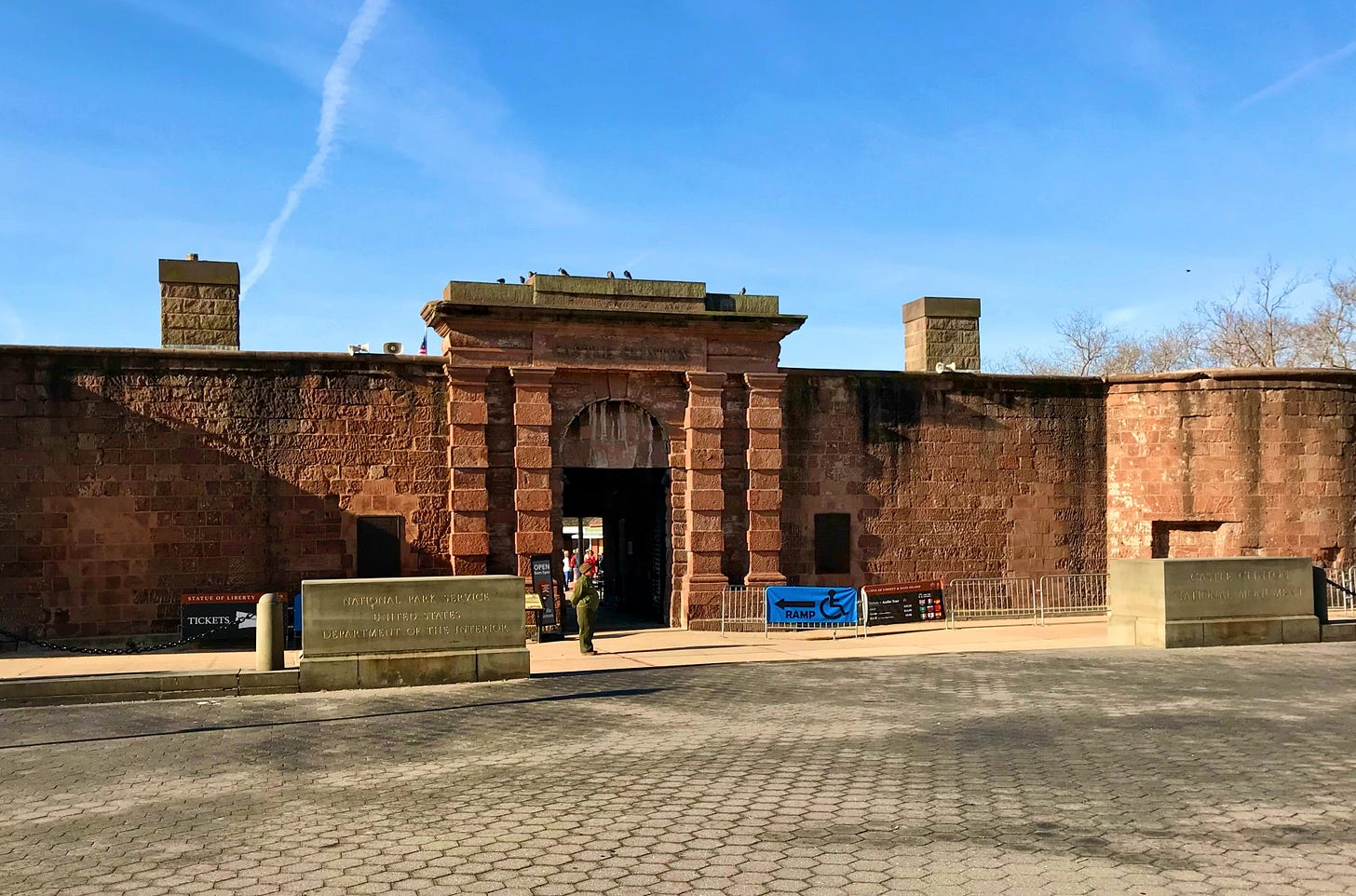 A large red brick fort. Over the stone arch, it is engraved CASTLE CLINTON. There is a stone slab at the gate that reads: National Park Service United States Department of the Interior