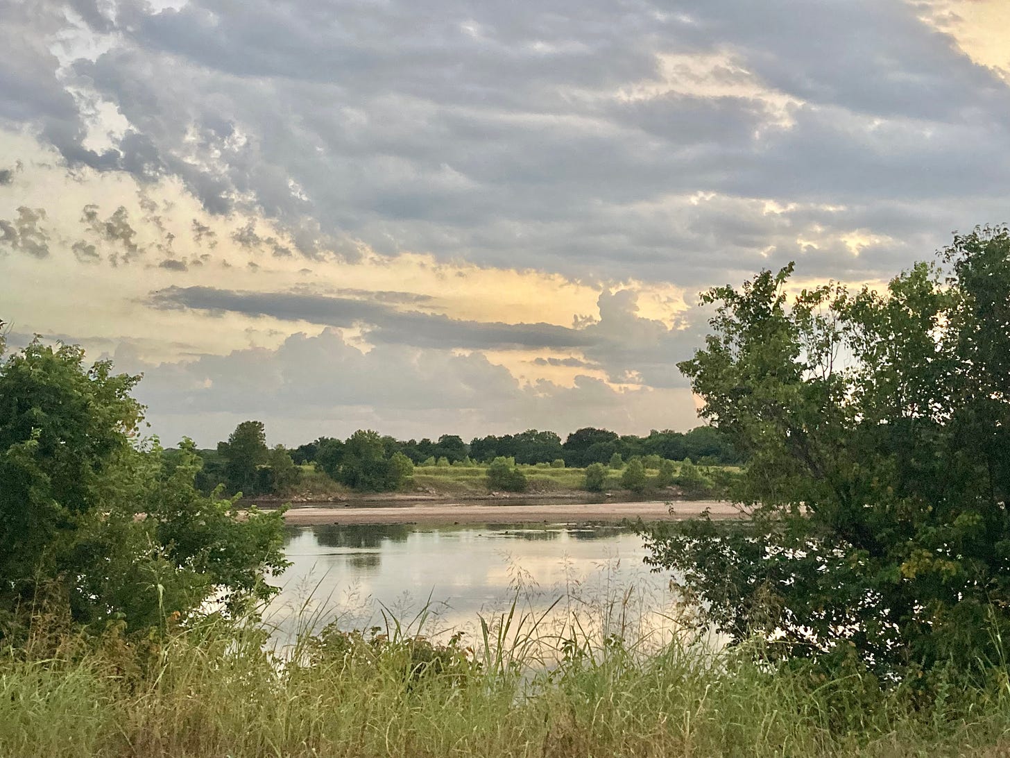 Framed by two trees and a blanket of grasses in the foreground, the photograph looks west over a placid Arkansas River beneath lavender grey clouds and patches of golden sky. The river is divided lengthwise by a long, pink sandbar and reflects layers of light and dark green vegetation on the opposite bank. 