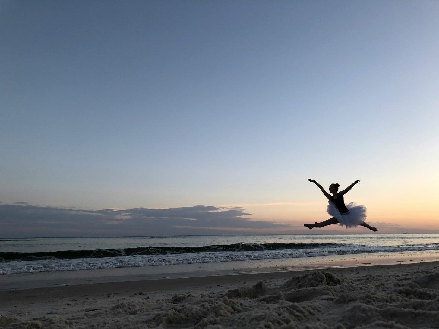 An evening beachside scene with a silhouetted ballerina in a tutu jumping over the water.